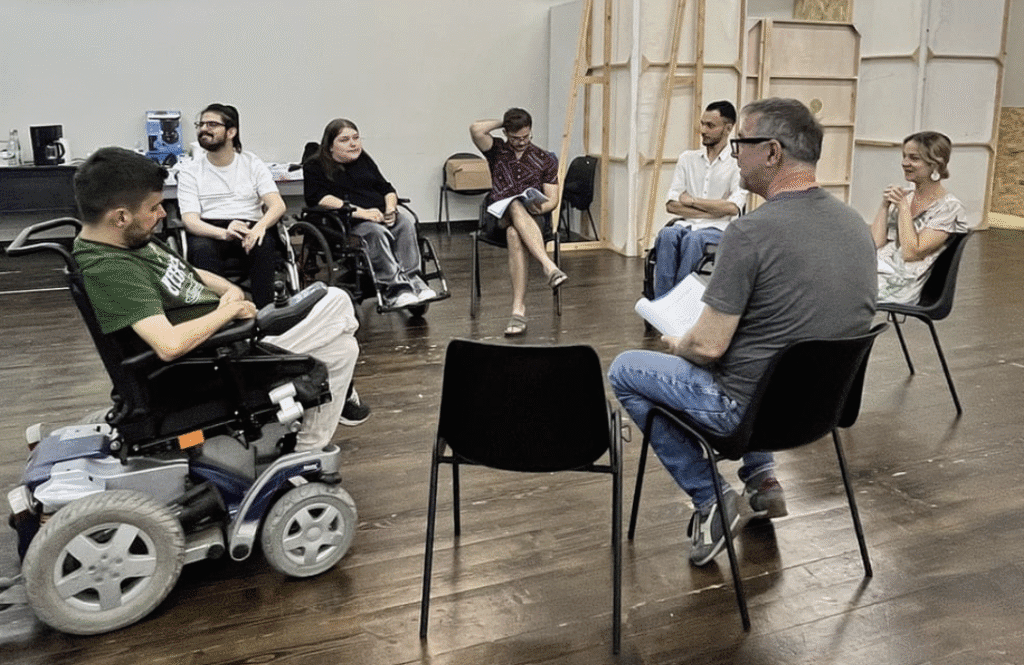 „O echipă de actori, dintre care mai mulți utilizatori de scaun rulant, participă la o repetiție într-o sală de teatru, așezați în cerc, discutând și citind din scenariu. Atmosfera este concentrată și prietenoasă.”
